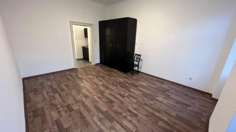 Heller Raum mit Holzplankenboden und einem schwarzen Kleiderschrank, der zu einer Kochnische führt.
