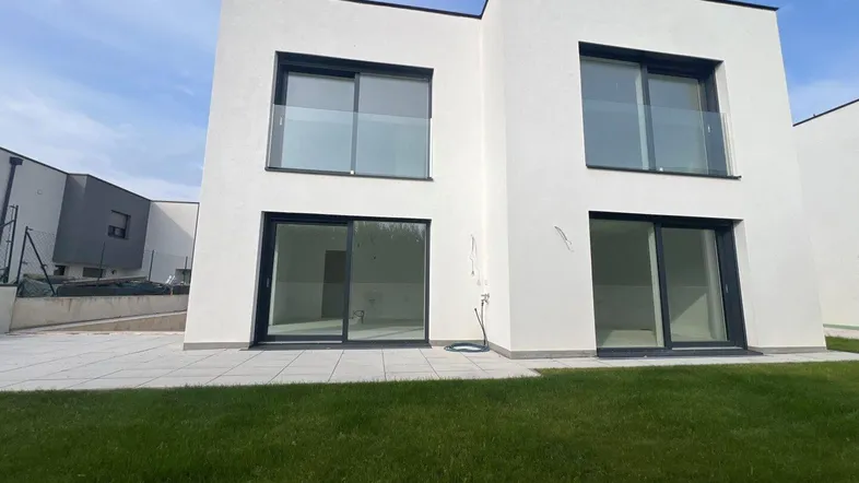 Moderne weiße Hausfassade mit großen Fenstern, Schiebetüren und einer kleinen Rasenfläche.
