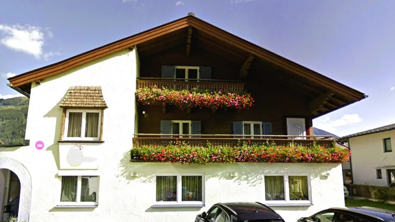 Traditionelles alpines Gebäude mit weißer Fassade, Holzbalkonen und blühenden Blumenkästen unter blauem Himmel.