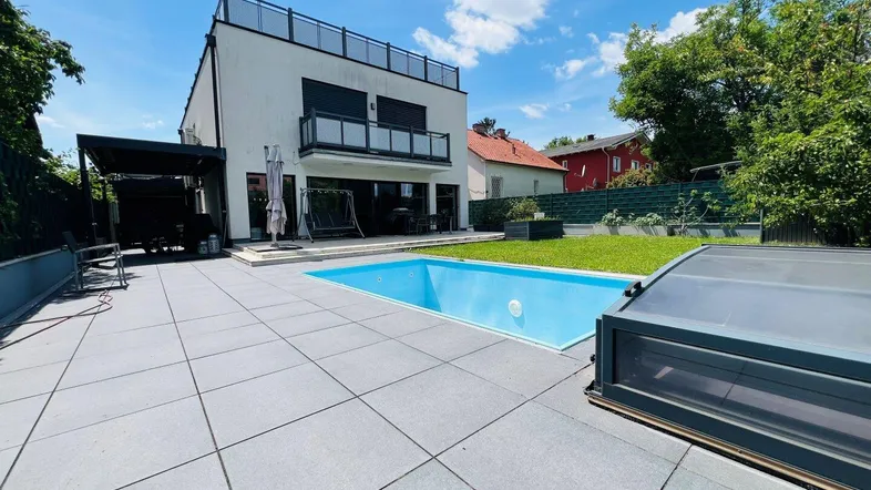 Modernes Haus mit Swimmingpool, großer Terrasse und Gartenbereich unter blauem Himmel.