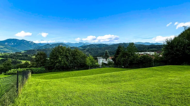 Weitläufige grüne Wiesen mit Blick auf die umliegende Berglandschaft unter blauem Himmel.