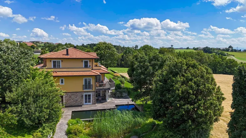 Haus mit Teich und viel Grün in ländlicher Umgebung unter blauem Himmel.