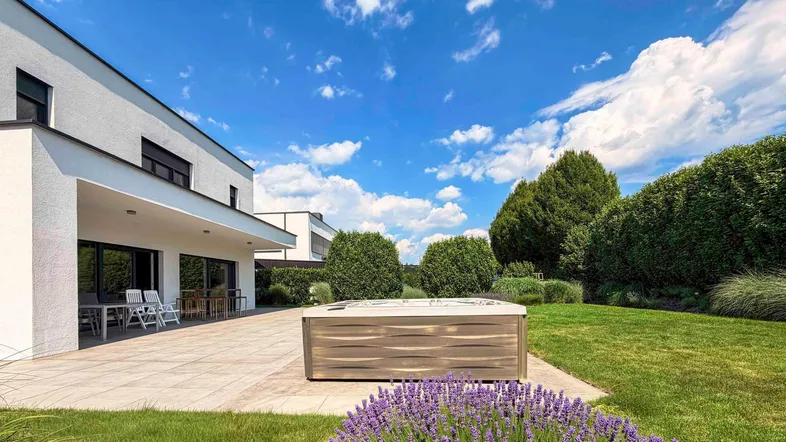 Moderner Gartenbereich mit großer Terrasse, Whirlpool und gepflegter Rasenfläche unter blauem Himmel.
