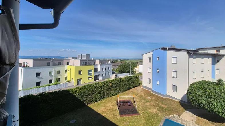 Blick von einem Balkon auf eine Wohnanlage mit Grünflächen, Spielplatz und Fernsicht.