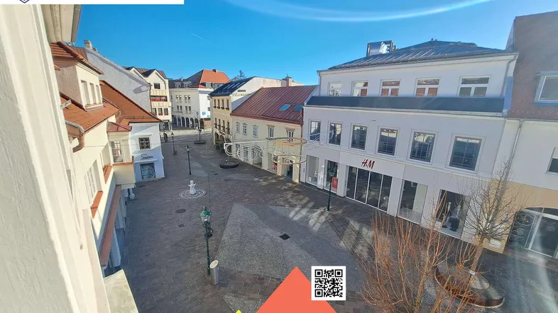 Blick auf einen belebten Stadtplatz mit Geschäften und historischen Gebäuden unter blauem Himmel.