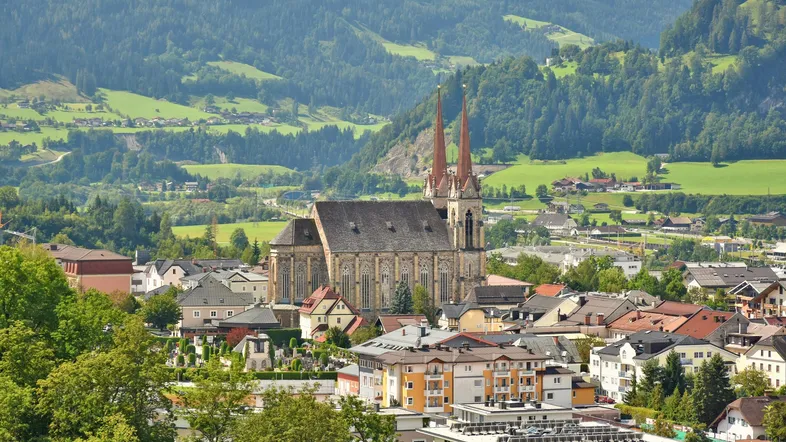 Panoramablick auf eine malerische Stadt mit einer imposanten Kirche, umgeben von grünen Hügeln und Bergen.