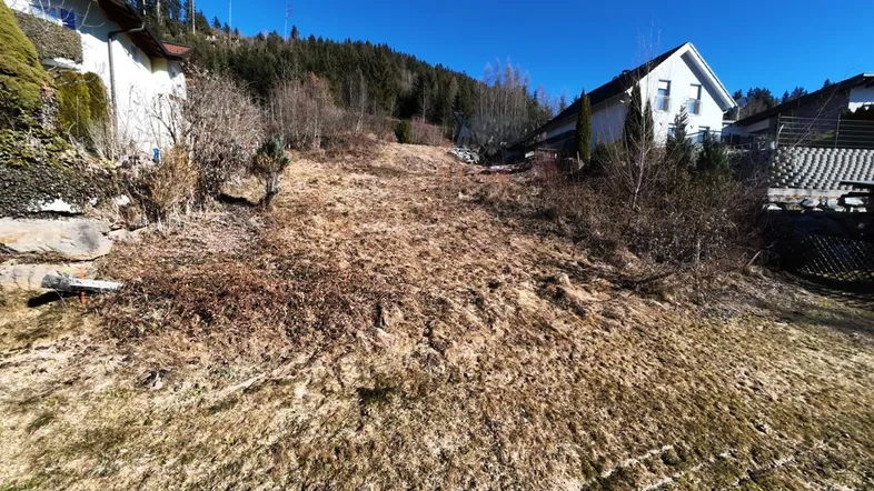 Ungenutztes Grundstück mit Blick auf umliegende Häuser und bewaldete Hügel unter blauem Himmel.
