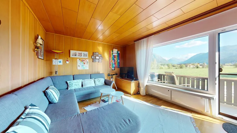 Helles Wohnzimmer mit Holzvertäfelung, großem Sofa und weitem Blick auf die Berge und grüne Landschaft.
