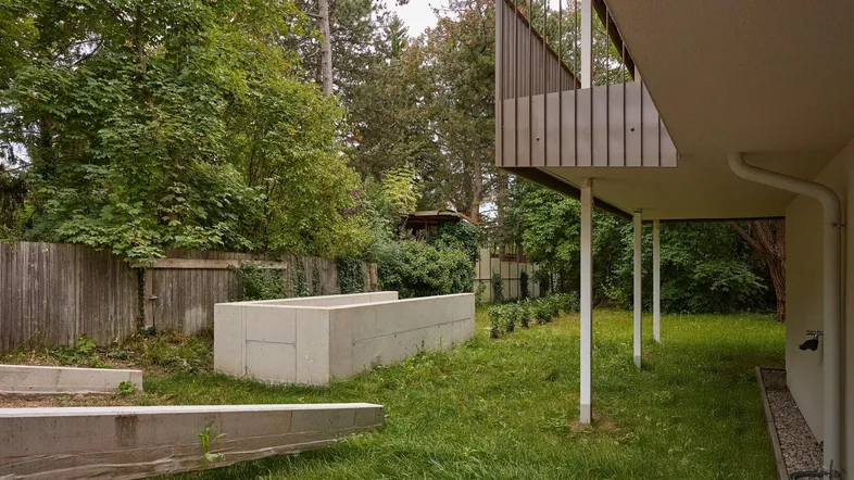 Grüner Gartenbereich mit Baumbestand und einem Balkon im Hintergrund des modernen Gebäudes.