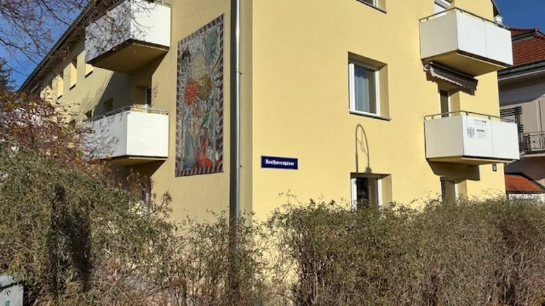 Gelbes Mehrfamilienhaus mit mehreren Balkonen und einem großen Wandgemälde an der Fassade.
