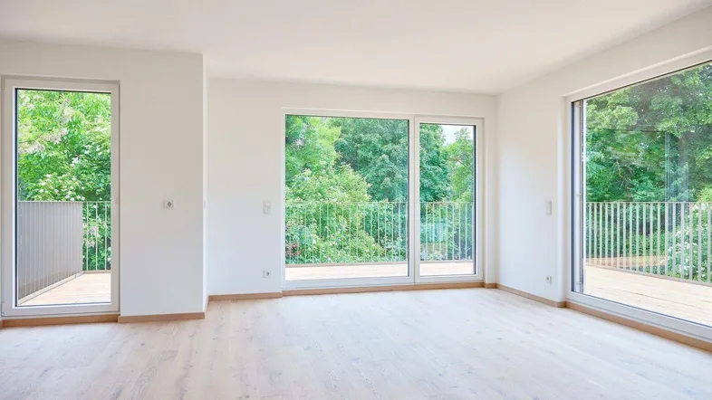Helles, leeres Zimmer mit Holzboden und Zugang zu mehreren Balkonen mit Blick ins Grüne.