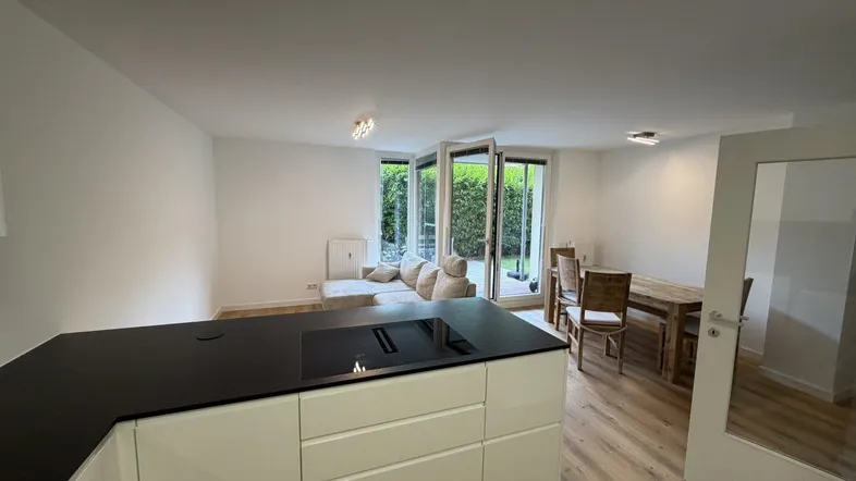 Heller offener Wohn-Essbereich mit moderner Küche, Laminatboden und Zugang zur Terrasse mit grüner Hecke.