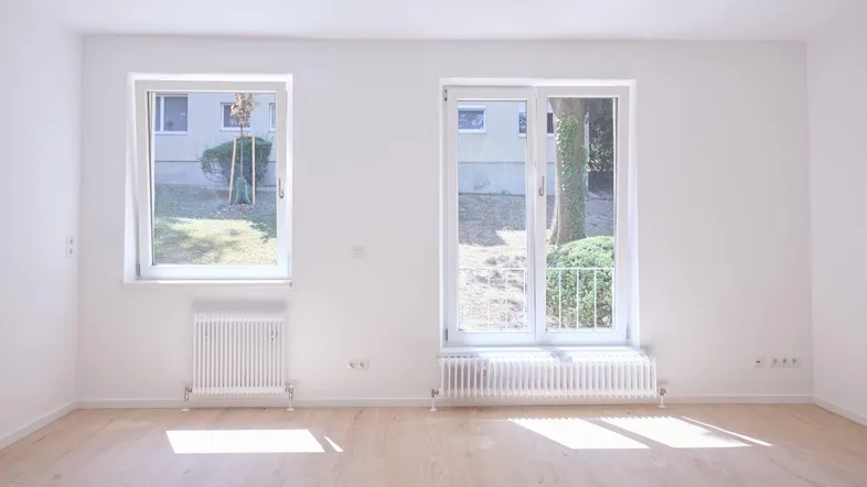 Helles, leeres Zimmer mit hellem Holzboden, weißen Wänden und großen Fenstern mit Blick ins Grüne.