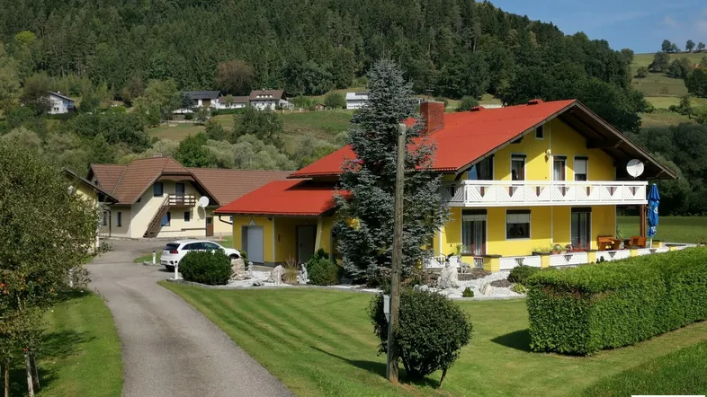 Gelbes Wohnhaus mit rotem Dach, umgeben von gepflegtem Rasen und bewaldeten Hügeln.