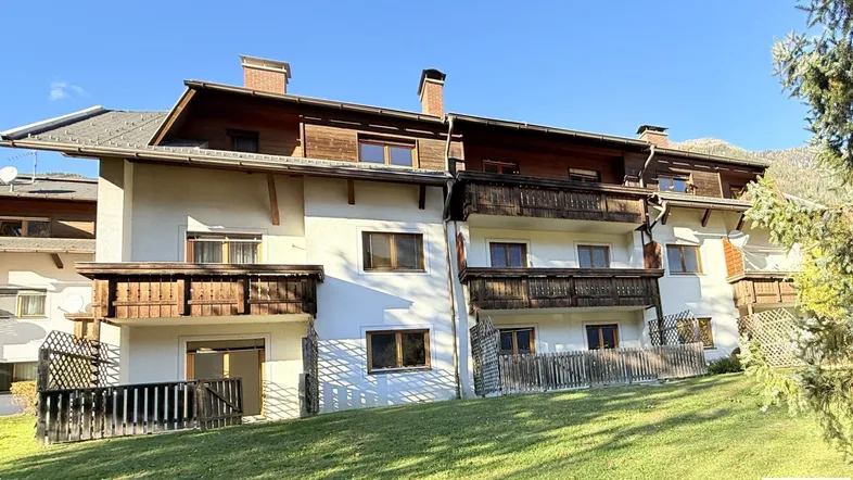 Mehrfamilienhaus mit traditionellen Holzbalkonen und gepflegter Rasenfläche in sonniger alpiner Umgebung.