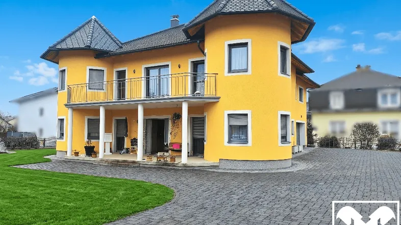 Gelbes Haus mit zwei Türmen, Balkon und gepflasterter Auffahrt unter blauem Himmel.