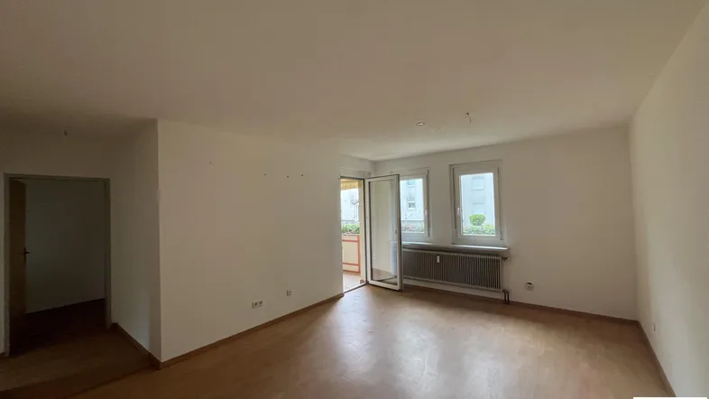 Helles, leeres Wohnzimmer mit Laminatboden und Zugang zu einem Balkon.