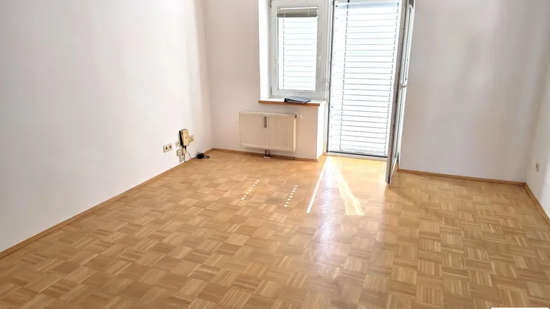 Unmöblierter Raum mit Parkettboden, weiß gestrichenen Wänden und Zugang zu einem Balkon.