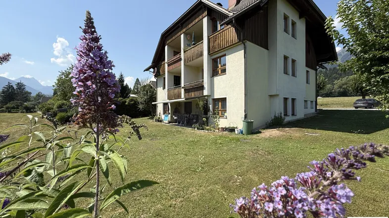 Mehrfamilienhaus mit mehreren Balkonen und einer Terrasse, umgeben von einem gepflegten Garten mit Bergblick.