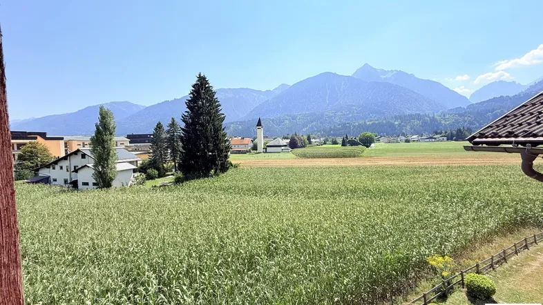 Weiter Blick über grüne Felder und Wiesen auf die umliegende Berglandschaft unter blauem Himmel.