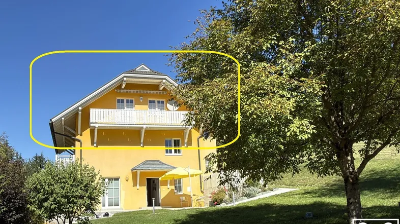 Gelbes Haus mit Balkonen und grüner Umgebung unter blauem Himmel.