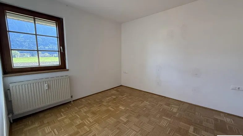 Helles Zimmer mit Parkettboden und Fensterblick ins Grüne, ausgestattet mit einem Heizkörper.