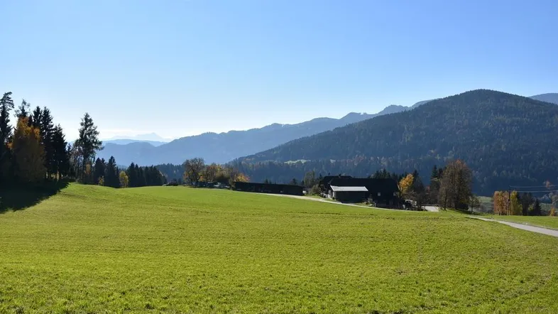 Weitläufige grüne Wiesenlandschaft mit einem Gebäude im Hintergrund und majestätischem Bergblick unter blauem Himmel.