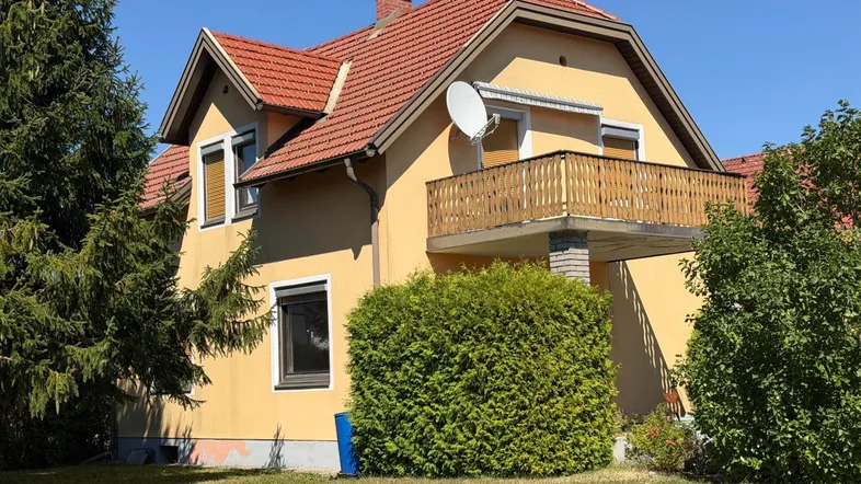 Gelbes Haus mit rotem Ziegeldach, Balkon und umgebender Grünfläche unter blauem Himmel.