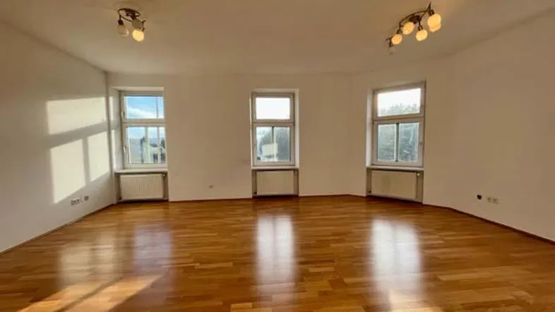 Helles, leeres Wohnzimmer mit Parkettboden und mehreren Fenstern, die viel Tageslicht hereinlassen.