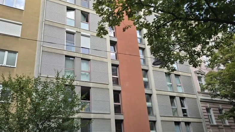 Moderne Wohnhausfassade mit vielen Fenstern und Jalousien, umgeben von Bäumen an einer Straße.