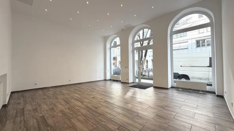 Weitläufiger Raum mit elegantem Holzfliesenboden, weißen Wänden und hohen Rundbogenfenstern zur Straße.