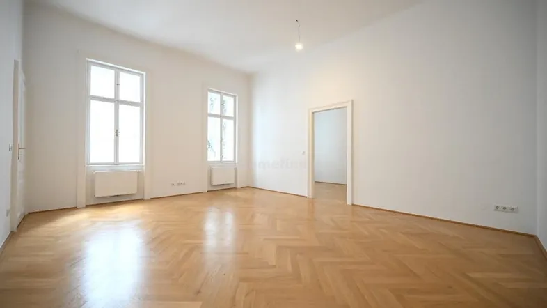 Helles, leeres Zimmer mit Fischgrätparkett und hohen Fenstern, ideal als Wohnbereich nutzbar.