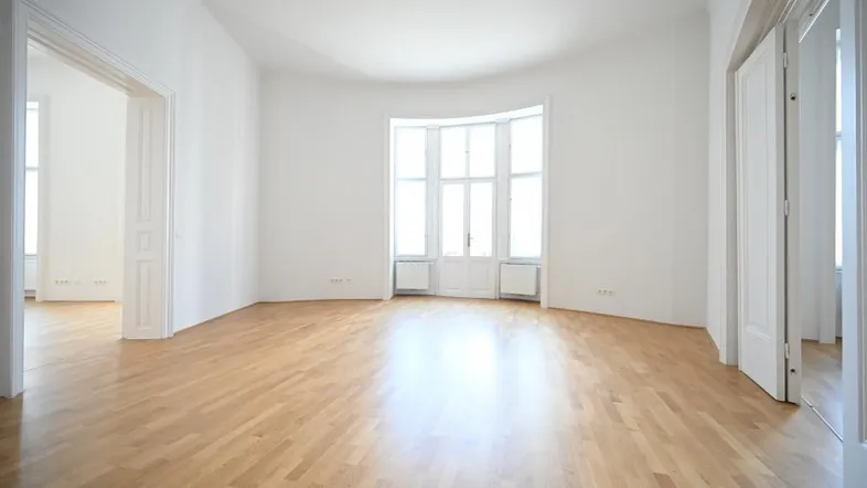 Geräumiges Wohnzimmer mit hellem Parkettboden und großen Fenstern, die viel Tageslicht hereinlassen.