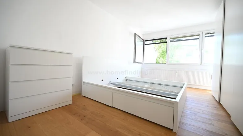 Helles Schlafzimmer mit Holzboden, großem Fenster und Stauraum unter dem Bett.