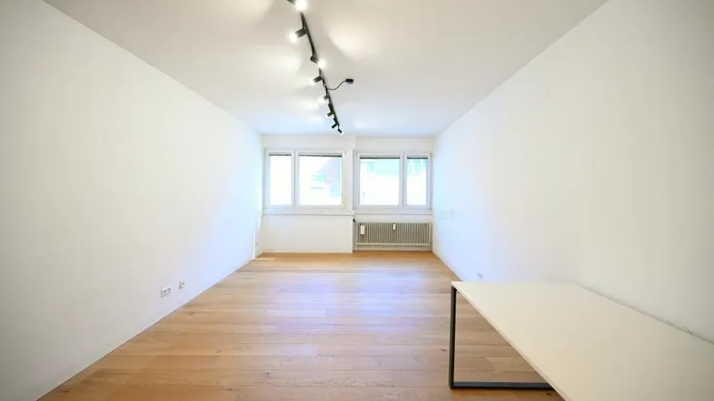 Helles Zimmer mit Holzfußboden, weißen Wänden und großen Fenstern, ausgestattet mit einer Schiene für Beleuchtung.