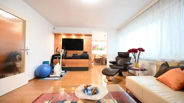 Helles Wohnzimmer mit beigem Sofa, Holzboden und einem Bereich für Fitnessgeräte.