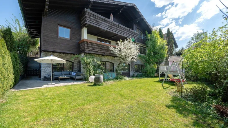 Großzügiger Gartenbereich mit Rasenfläche, Terrasse und Spielplatz vor einem traditionellen Holzhaus.