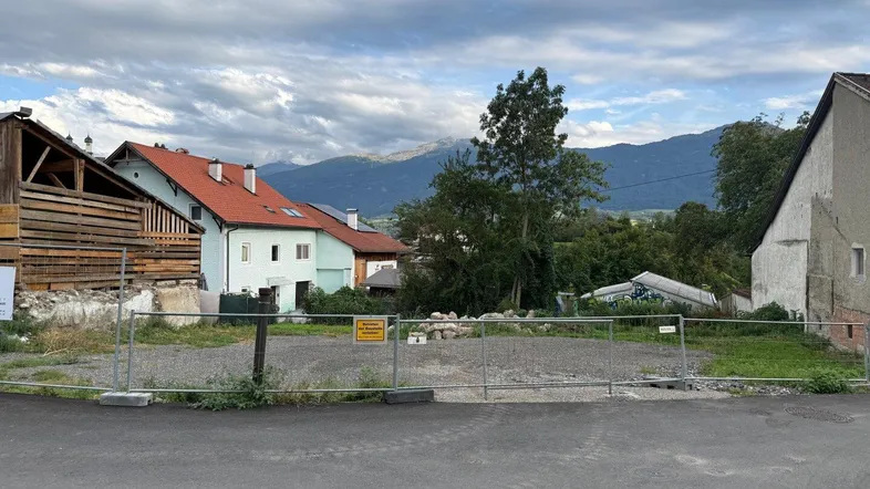 Leeres Grundstück mit Bauzaun, umgeben von Häusern und Bergen im Hintergrund unter bewölktem Himmel.