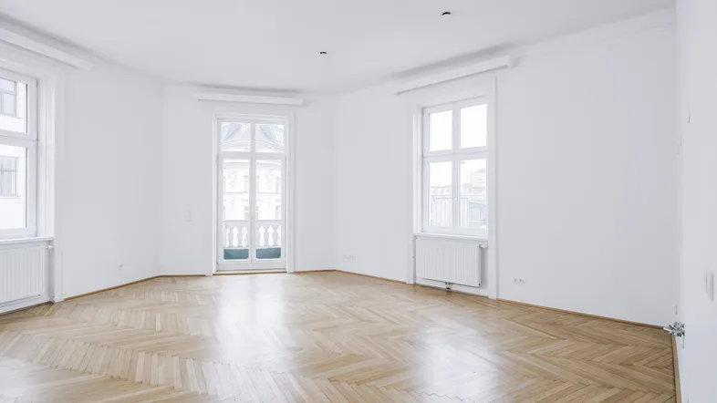 Helles, leeres Zimmer mit Fischgrätparkett, hohen Decken und Zugang zu einem französischen Balkon.