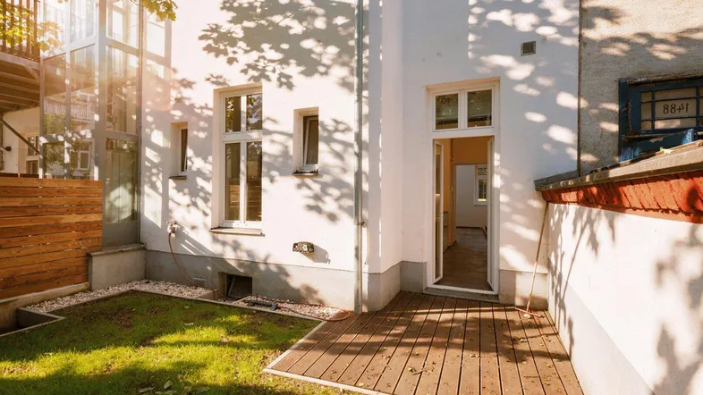 Kleine Terrasse mit Holzdeck und angrenzendem Rasenbereich, umgeben von Gebäuden im Hinterhof.