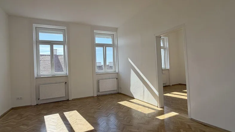 Helles Zimmer mit Fischgrätparkettboden, weißen Wänden und viel Tageslicht durch zwei Fenster.