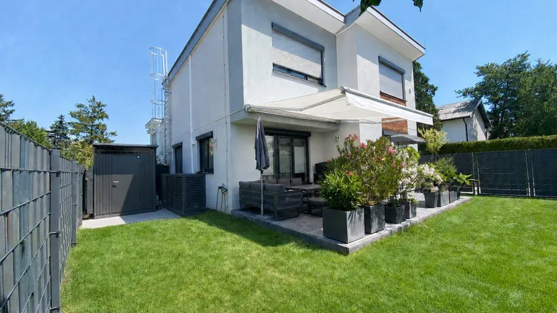 Modernes Haus mit Garten, Terrasse und Geräteschuppen unter blauem Himmel.