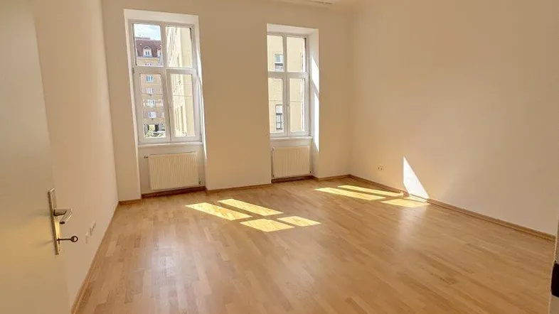 Helles Zimmer mit Parkettboden und zwei Fenstern, durch die viel Sonnenlicht einfällt.