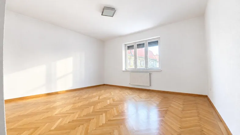 Helles, leeres Zimmer mit Fischgrätparkett und Fenster, das viel Tageslicht hereinlässt.