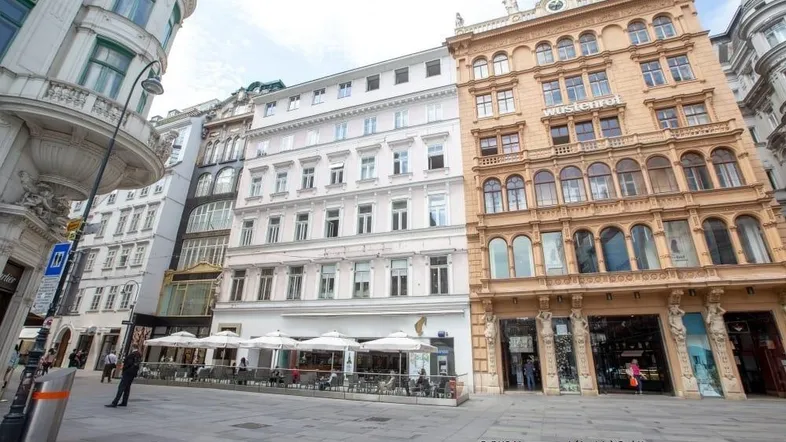 Historische Gebäudefronten an einer belebten Straße mit Außengastronomie und Passanten in der Stadt.
