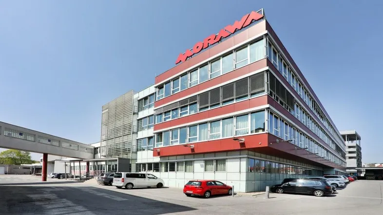 Modernes Bürogebäude mit roter Fassade, großen Fenstern und Parkplätzen im Freien unter blauem Himmel.