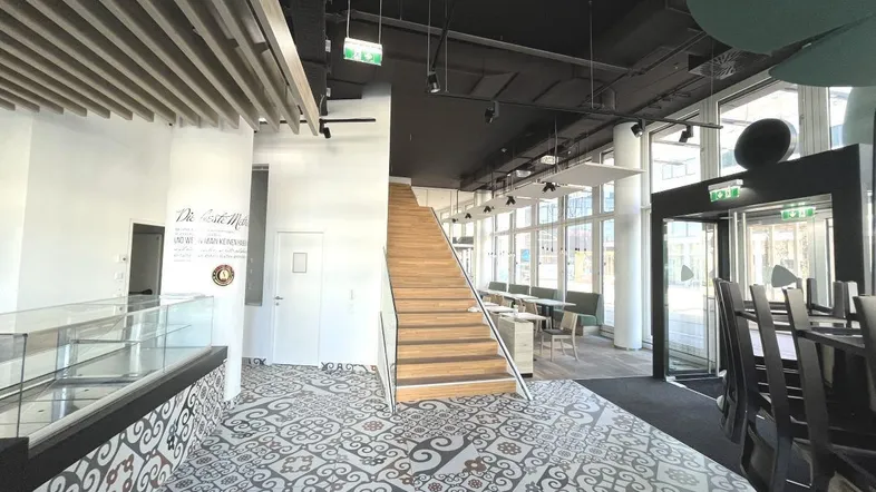 Geräumiger Innenbereich eines Cafés mit dekorativen Fliesen, Holztreppe und großen Fenstern.