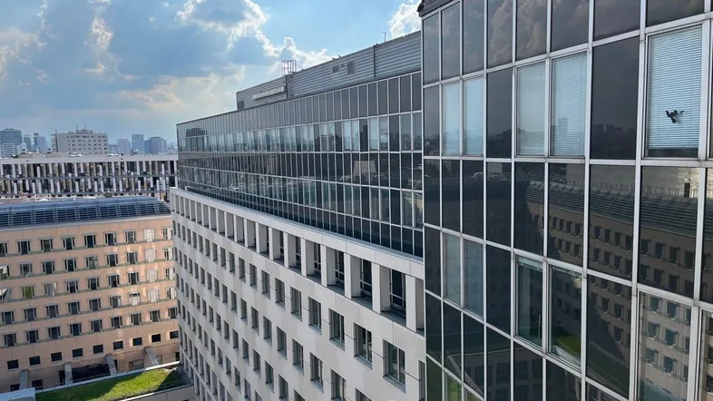 Blick auf die moderne Glasfassade eines Bürogebäudes mit weiteren Gebäuden und bewölktem Himmel im Hintergrund.