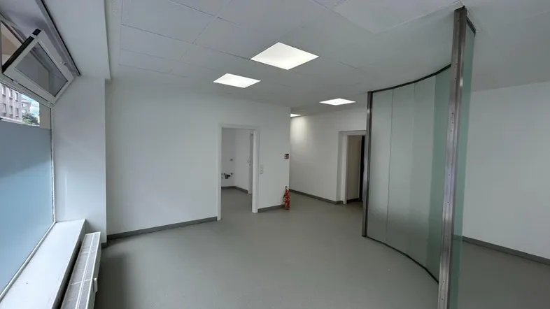 Heller Büroraum mit modernen Deckenleuchten, großen Fenstern und flexiblen Glaswänden für eine offene Gestaltung.