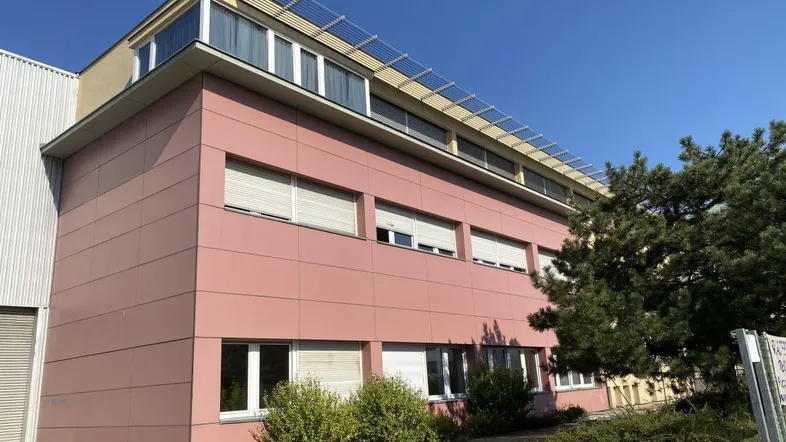 Gebäudeansicht mit rosafarbener Fassade, vielen Fenstern und Sonnenschutzlamellen unter blauem Himmel.
